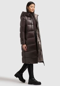 Brown puffer coat with a hood, featuring quilted texture, front zip, and side pockets. Worn over a dark top and paired with knee-high boots.