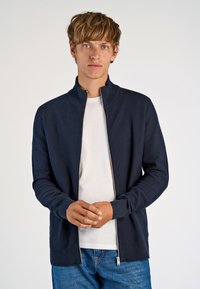 Marinblå zipjacka med hög krage och texturerad tyg. Matchad med en vit skjorta. Har ribbade ärmslut och nederkant. Enkel, modern design.