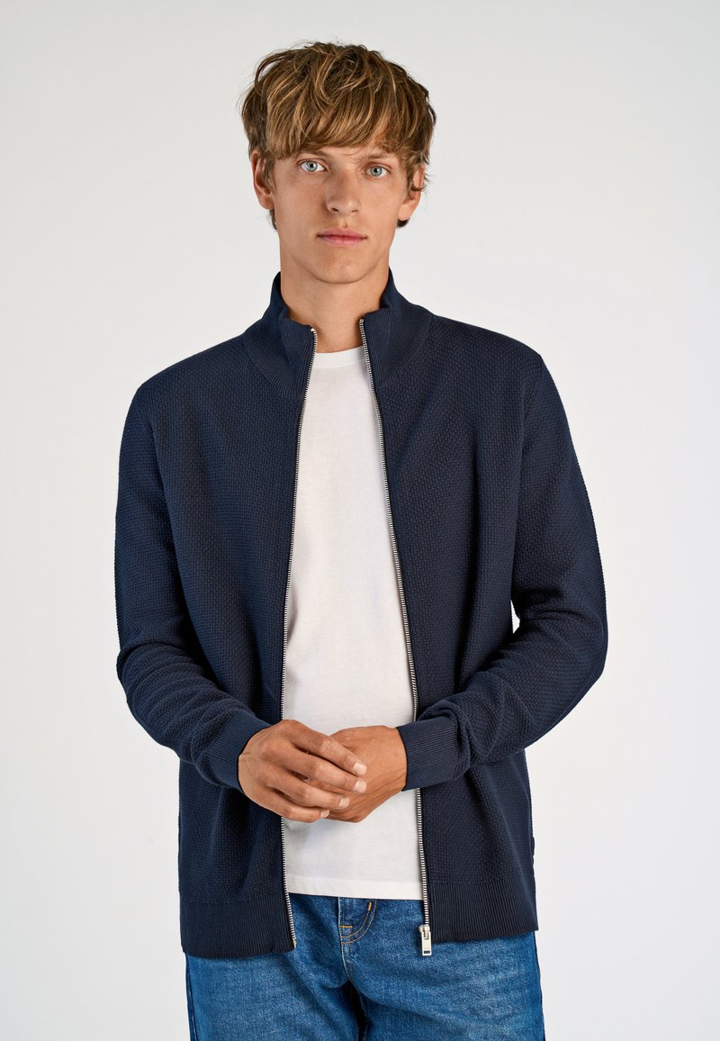 Marinblå zipjacka med hög krage och texturerad tyg. Matchad med en vit skjorta. Har ribbade ärmslut och nederkant. Enkel, modern design.