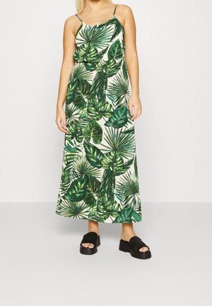 Woman wearing a sleeveless maxi dress with green tropical leaf print and black platform sandals standing against a plain background.