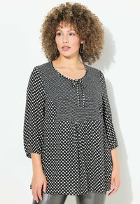 Haut noir et blanc à pois avec un col rond, des manches 3/4, un détail à nouer à l'avant et une conception froncée à la taille.