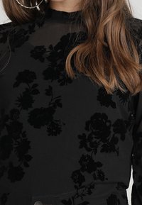 Svart topp med genomskinligt tyg och strukturerat blommönster. Mock neck-design och lång ärm. Accentuerad med stora silverfärgade hoops. 