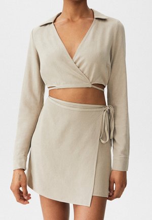 Crop top beige style cache-cœur à manches longues, associé à une mini-jupe portefeuille assortie nouée à la taille.