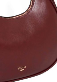 Burgundy leather bag with a smooth texture, curved shape, gold zipper detailing, and gold "Dune" branding on the front.