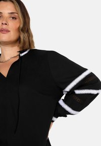 Schwarze Bluse mit V-Ausschnitt, versehen mit weißer Stickerei an den Ärmeln und elastischen Manschettendetails. Strukturierter Stoff und lockere Passform.