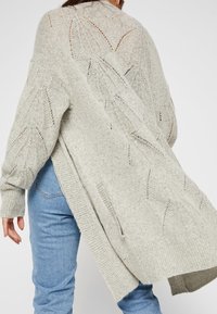 Woman wearing light grey knit cardigan with leaf patterns and light blue jeans, shown from the back against a plain background.