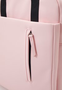 Gros plan sur un sac rose clair avec une fermeture éclair noire et une languette sur la poche avant, ainsi que des poignées noires s'étendant vers le haut.