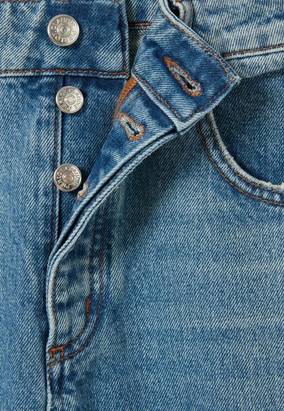 Giacca in denim blu chiaro con bottoni a pressione argentati, cuciture a vista e una leggera texture usurata. Vista ravvicinata dei dettagli del tessuto.