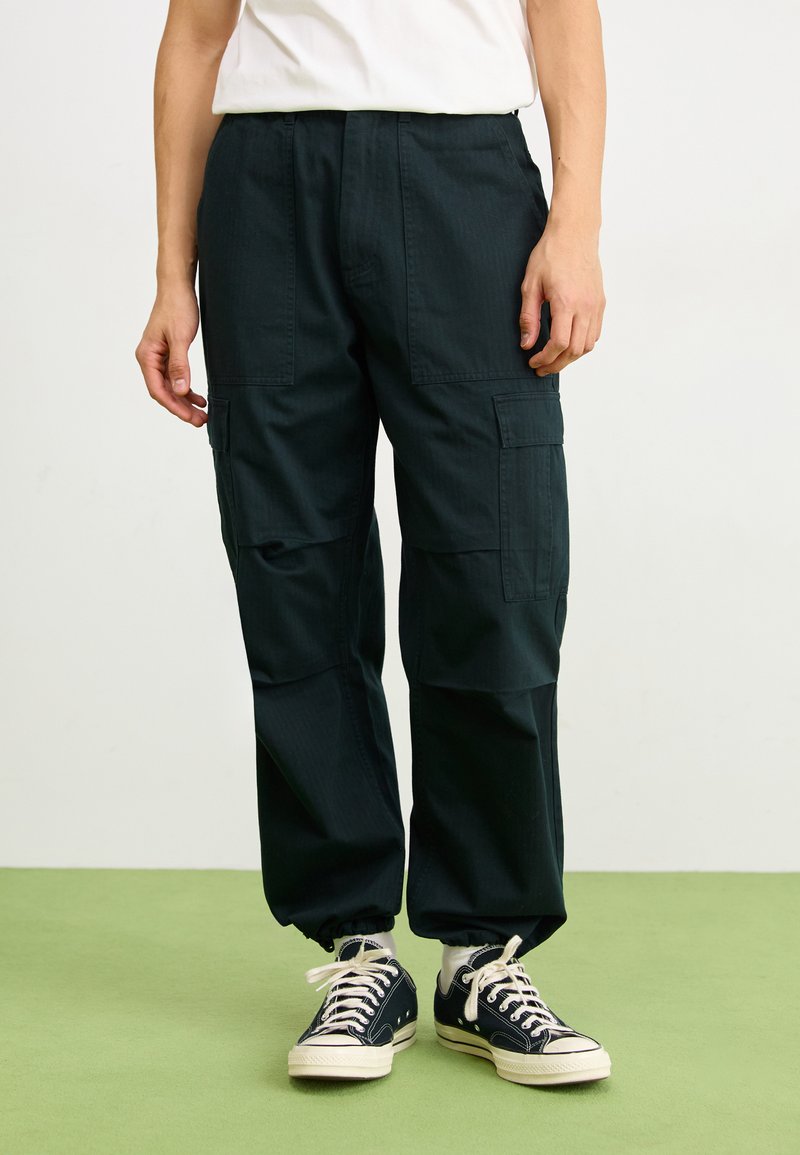 Pantalones cargo de color verde oscuro con múltiples bolsillos laterales, puños elásticos y un corte relajado, combinados con zapatillas negras y calcetines blancos.