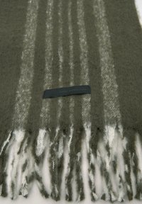 Olivnozeleni šal iz mehkega materiala, s belimi črtami in resicami na koncih. Črni napis na etiketi glasi: "KLASIČNO. MODERNO. TVOJE."