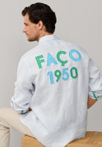 Man zittend, draagt een lichtblauw-wit gestreept shirt met groen-blauwe letters "FAÇO 1950" op de achterkant.