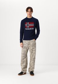 Marineblauer Strickpullover mit einem rot-weißen norwegischen Flaggenmuster, kombiniert mit hellgrauen gemusterten Hosen und schwarzen Turnschuhen.