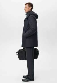 Parka impermeabile blu navy con cappuccio, caratterizzata da una vestibilità comoda e tasche laterali, abbinata a pantaloni grigi su misura e scarpe in pelle nere.