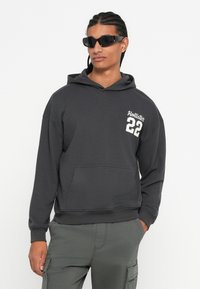 Άνδρας που φορά μαύρα γυαλιά ηλίου, σκοτεινές γκρι μπλούζες Hollister με "22" στο στήθος και γκρι cargo παντελόνια, στέκεται με το ένα χέρι στην τσέπη.