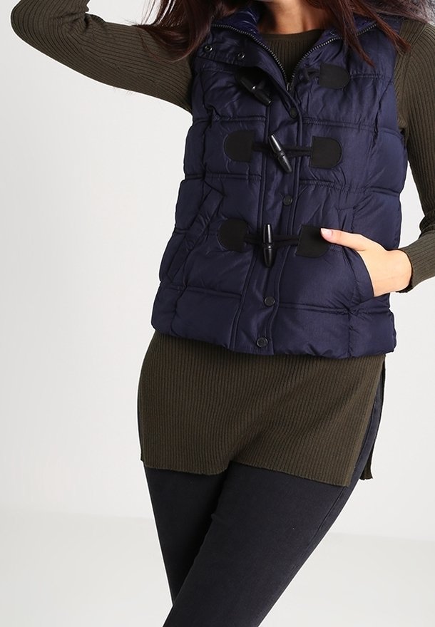 Femme portant un gilet matelassé bleu marine avec boutons à bascule sur un haut à manches longues côtelé olive et un pantalon noir, posant avec une main dans la poche.