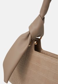 Tan faux läderhandväska med krokodilstruktur, guldfärgade öglor och ett knutet handtag på en vit bakgrund.