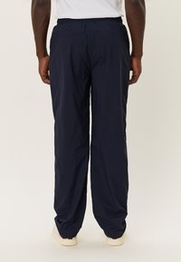 Osoba v volných tmavě modrých kalhotách, bílých teniskách a bílém tričku, stojící zády k fotoaparátu.