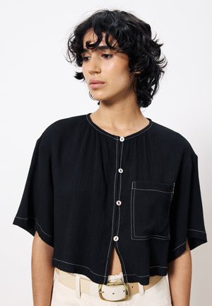 Chemise noire raccourcie à manches courtes, en tissu texturé, avec fermeture à boutons présentant des coutures contrastantes, et une poche sur le sein gauche.
