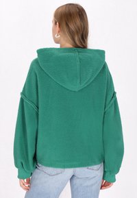 Grøn fleece hoodie med hætte, lange ærmer og en tekstureret overflade. Har en afslappet pasform med sænkede skuldre og ribbede manchetter.