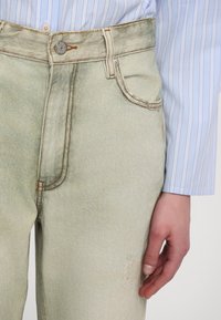 Gros plan sur un pantalon en jean beige avec des coutures visibles, un bouton en métal et une main reposant sur le côté portant une chemise rayée bleue et blanche.