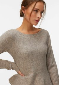 Femme aux cheveux châtain clair attachés en un chignon bas, portant des boucles d'oreilles argentées en forme de créoles et un pull en maille marron clair, regardant sur le côté avec une main sur la taille.