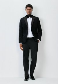 Schwarzer Samt-Sakko mit Schalkragen, weißes Hemd, schwarze Fliege und schwarze Hose mit glänzenden schwarzen Schuhen.