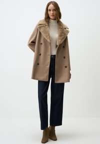 Cappotto doppiopetto in lana marrone chiaro con colletto in pelliccia sintetica, dotato di grandi bottoni e una vestibilità morbida, abbinato a jeans scuri e stivaletti beige.