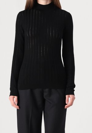 Femme portant un haut noir moulant côtelé à manches longues avec col montant, associé à un pantalon noir ample sur un fond uni.