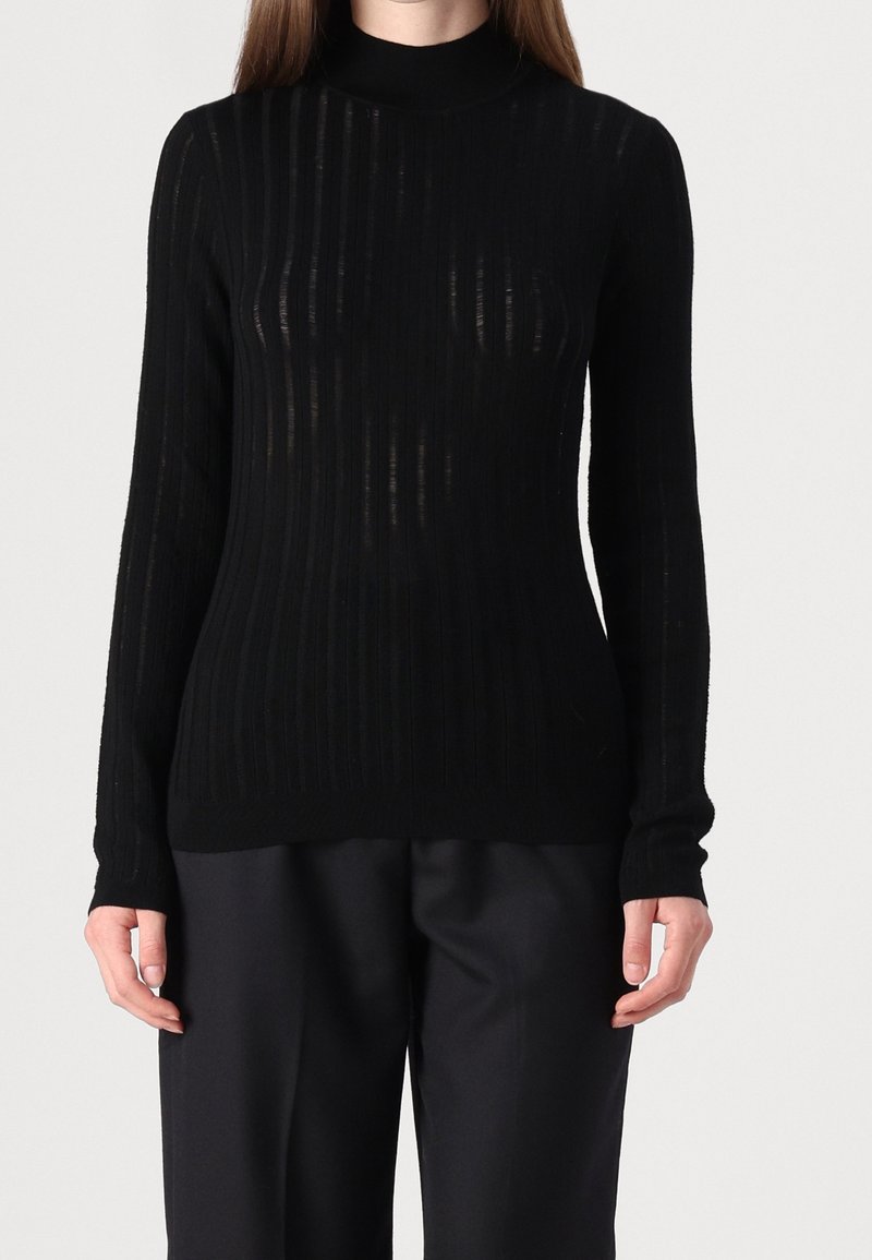 Femme portant un haut noir moulant côtelé à manches longues avec col montant, associé à un pantalon noir ample sur un fond uni.