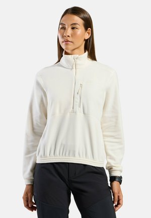 Frau, die eine weiße Fleecejacke mit halbem Reißverschluss und schwarze Hose trägt, steht vor einem schlichten Hintergrund und schaut leicht zur Seite.