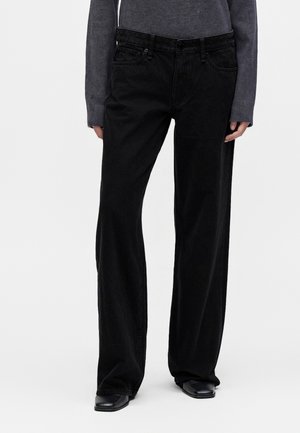 Personne portant un pantalon noir ample, un haut gris à manches longues et des chaussures noires, debout devant un fond blanc uni.