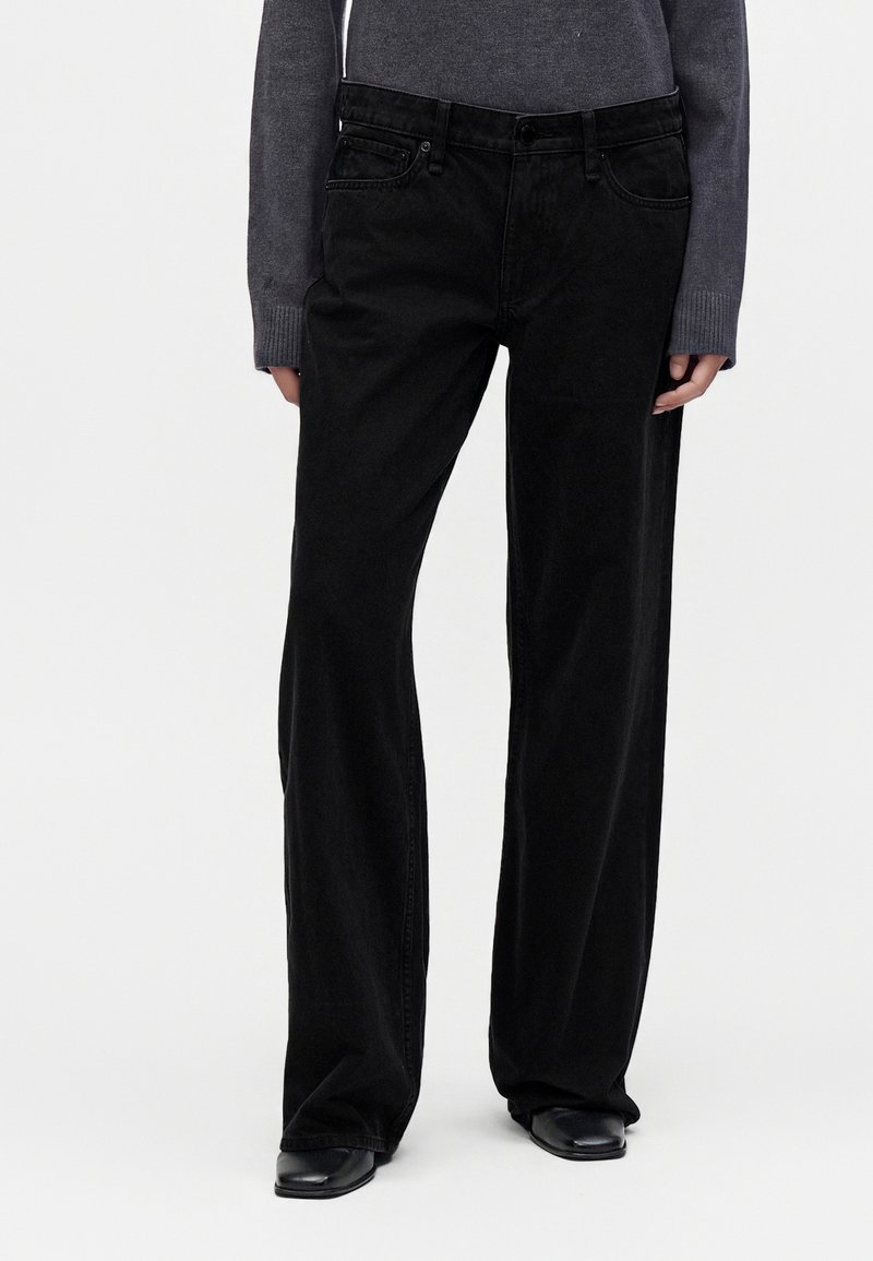 Personne portant un pantalon noir ample, un haut gris à manches longues et des chaussures noires, debout devant un fond blanc uni.