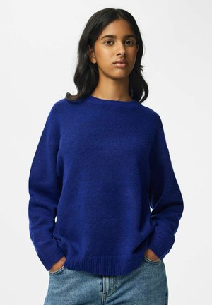 Jeune femme aux longs cheveux bruns portant un pull bleu et un jean bleu clair, les mains dans les poches, sur un fond blanc uni.