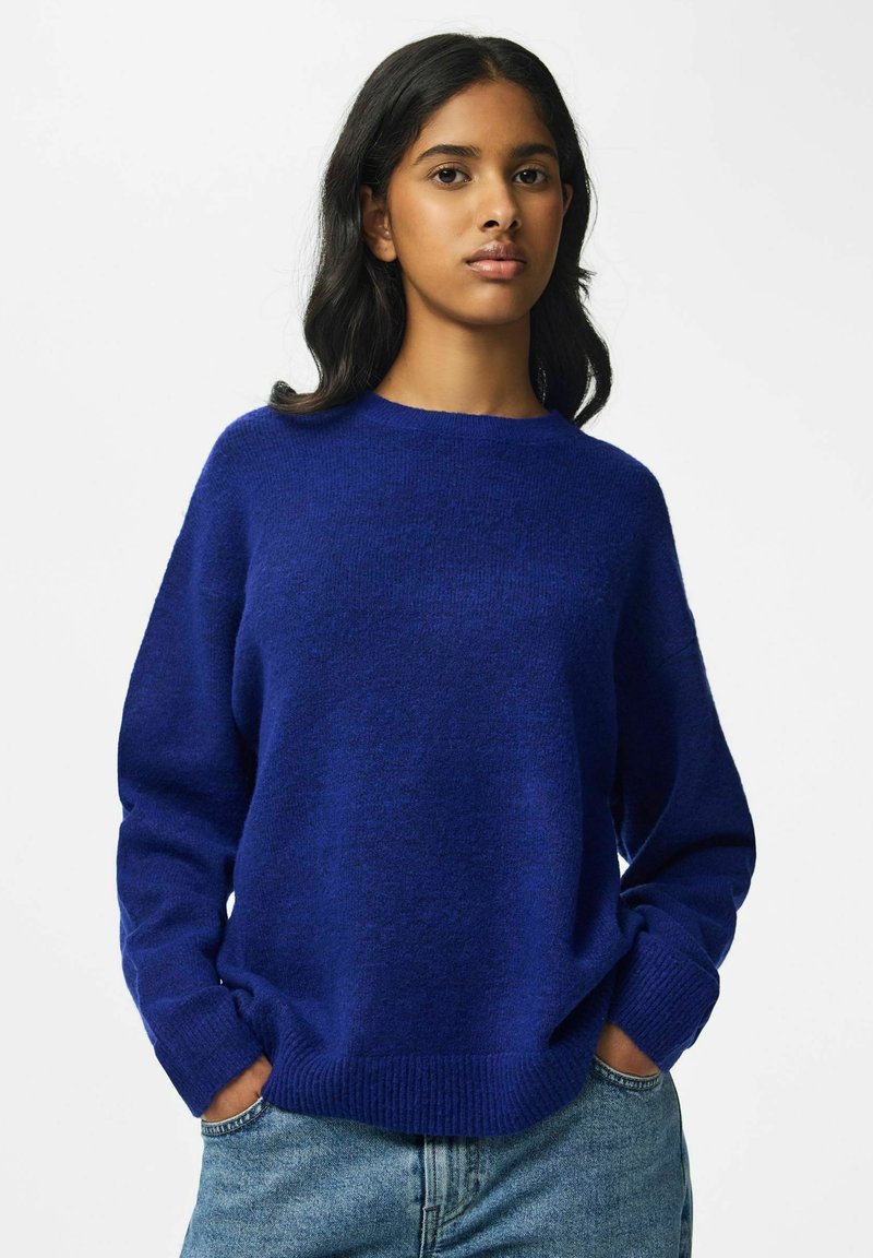 Jeune femme aux longs cheveux bruns portant un pull bleu et un jean bleu clair, les mains dans les poches, sur un fond blanc uni.