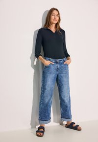 Musta pitkähiha polo-paita, siniset leveät farkut vaalealla pesulla ja käännetyillä lahkeilla, tummansiniset liukumattomat sandaalit, joissa on kaksinkertaiset soljet.