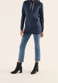 Námořnická mikina s kapucí na stahování, bočními kapsami a volnějším střihem, ve spojení s světle modrými džíny a černými kotníkovými botami.