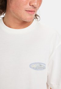 Hvid bomulds t-shirt med et lyseblåt broderet logo på venstre brystområde. Standard rund hals design med korte ærmer.