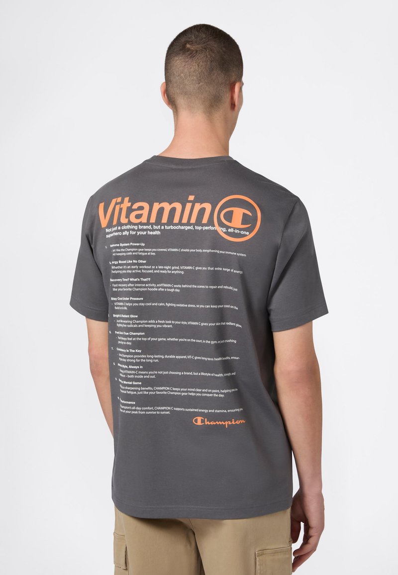 Młoda osoba stoi tyłem, ubrana w ciemnoszary t-shirt z dużym pomarańczowym napisem "Witamina C" oraz szczegółowym białym tekstem na plecach, w beżowych spodniach.