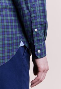 Grön och blå rutiga långärmade skjorta med knäppta manschetter, rundad nederkant och textilstruktur. Blå undertröja synlig nedanför.