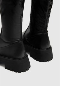 Bottes noires montantes jusqu'au genou avec une tige en cuir lisse, une semelle en caoutchouc rainurée proéminente et une fermeture éclair latérale. Design minimaliste sans motifs.