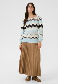 Femme portant un pull zigzag bleu clair, blanc, noir et marron avec une longue jupe plissée marron et des baskets marron.