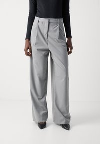 Pantalons gris ajustés avec une coupe ample, devant plissé et texture lisse. Portés avec un haut noir ajusté et des talons noirs pointus.