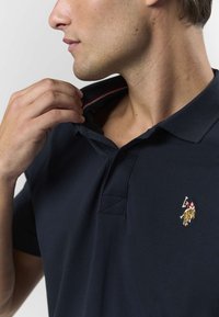 Polo bleu marine avec un col de couleur contrastante, orné d'un petit logo brodé. Le tissu semble lisse et légèrement texturé.