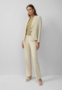 Beige tailored blazer gecombineerd met bijpassende trousers, over een saliegroen top met subtiele stippenpatronen. Witte, strap-sandalen maken de look compleet.