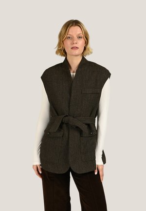 Femme portant une veste sans manches texturée marron foncé avec des épaulettes, ceinture nouée à la taille, par-dessus un haut blanc à manches longues et un pantalon foncé.