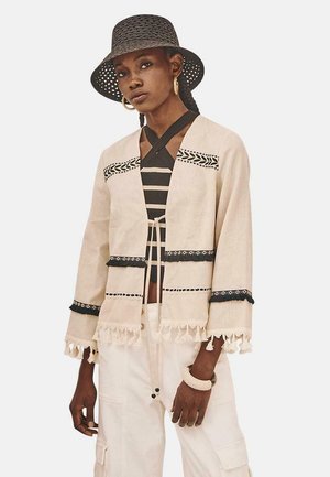 Chaqueta beige con detalles negros bordados, dobladillo con borlas y abertura frontal. Combinada con una blusa negra a rayas y pantalones anchos color crema.