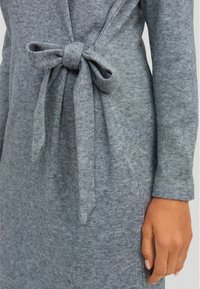Abito grigio a maglia con design avvolgente, vita legata, maniche lunghe e una texture morbida. Il tessuto presenta un sottile motivo mélange.
