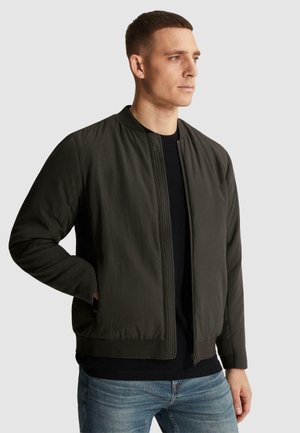 Mann trägt eine dunkelgrüne Bomberjacke über einem schwarzen Shirt und blauen Jeans, steht mit einer Hand in der Tasche vor einem einfarbigen Hintergrund.