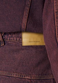 Dark burgundy denim garment with contrasting stitching, featuring a leather patch with embossed branding at the waistband.