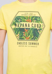 T-shirt en coton jaune avec un graphique hexagonal présentant des plantes tropicales et le texte "HAVANE CUBA" et "ÉTÉ SANS FIN" en noir.
